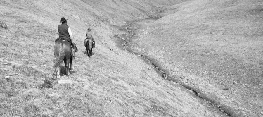 Black and white photo of two people riding horses in mountains